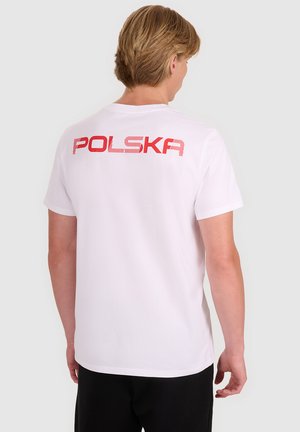 Osoba o jasnobrązowych włosach ubrana w białą koszulkę z czerwonym napisem "POLSKA" na górnej części pleców, stojąca na jednolitym jasnym tle.