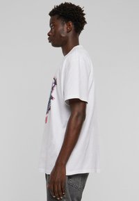 Hvid bomulds t-shirt med korte ærmer, der har et farverigt grafisk design på forsiden og en afslappet pasform. Sidevisning viser rund halsudskæring.
