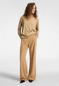 Donna con capelli ricci che indossa un maglione beige e pantaloni a gamba larga color cammello, con le mani nelle tasche, che guarda di lato su uno sfondo neutro.