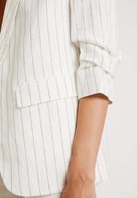 Blazer blanc à rayures verticales en gris clair, présentant un col cranté, une poche unique et un détail de manches roulées. Tissu doux et texturé.