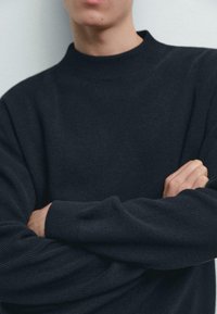 Ciemnoszary dzianinowy sweter z wysokim kołnierzem, prążkowaną fakturą i długimi rękawami. Ręce skrzyżowane, prezentujące wzór i design tkaniny.