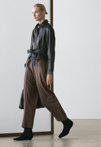 Kvinde går indendørs iført en sort læderjakke med bælte, brune løse bukser og sorte ankelstøvler, bærende på en sort håndtaske.