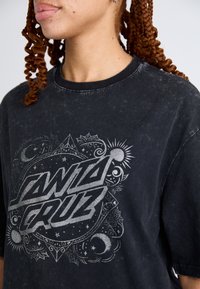 Tricou din bumbac negru cu un print grafic gri care prezintă elemente de design intricate, inclusiv motive cu stele, soare și lună. Decolteu rotund.