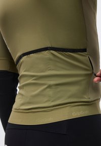 Κοντινό πλάνο ενός ατόμου με ποδηλατικό μπλουζάκι ελιάς που ξεκουμπώνει μια πίσω τσέπη με μαύρη επένδυση, φορώντας μαύρα ποδηλατικά σορτς από κάτω.