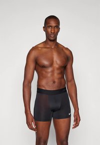 Moški brez srajce, oblečen v črne bokserice Nike, stoji ob preprostem svetlem ozadju, obrnjeno naprej z rokami sproščeno ob straneh.