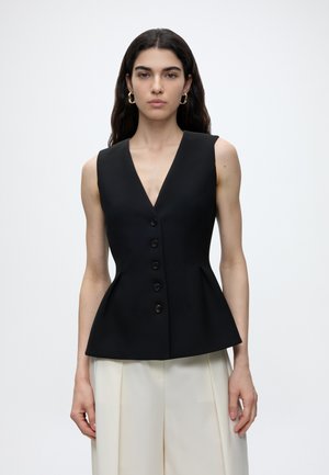 Femme aux longs cheveux foncés porte un gilet noir sans manches boutonné et un pantalon blanc à jambes larges sur un fond clair uni.