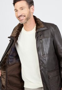 Man die glimlacht terwijl hij een donkerbruin leren jack met bontvoering draagt over een wit gebreid sweater.