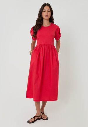 Mujer de pie vistiendo un vestido midi rojo brillante con mangas cortas abullonadas, cintura fruncida, bolsillos laterales y sandalias planas negras con tiras.