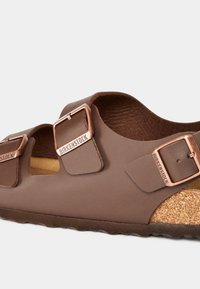 Sandali Birkenstock marroni con due cinghie regolabili, accessori in oro rosa, soletta in sughero e upper in sintetico testurizzato. Suola in gomma antiscivolo.