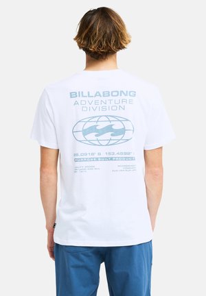 Persoon met lichtbruin haar die een wit Billabong Adventure Division T-shirt en een blauwe broek draagt, gezien van achteren.
