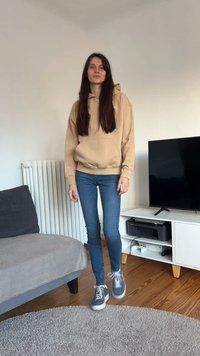 Rudos spalvos džemperis su gaubtu, mėlyni džinsai ir raštuoti sportbačiai. Stovi ant šviesios kiliminės dangos šviesiame kambaryje, kuriame yra pilkas sofa ir televizorius.