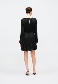 Vrouw met haar in een knot, gekleed in een zwarte, zwart, lange mouwen geplooide jurk met een gerafelde zoom en een kettingriem, staand met haar rug naar voren tegen een witte achtergrond.