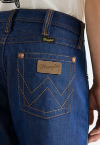 Τα σκούρα μπλε τζιν από τζιν ύφασμα διαθέτουν ένα χαρακτηριστικό μοτίβο ραφής ζιγκ-ζαγκ στην πίσω τσέπη, με ένα δερμάτινο έμβλημα λογότυπου και μεταλλικά μέρη σε χαλκοκόκκινο χρώμα.