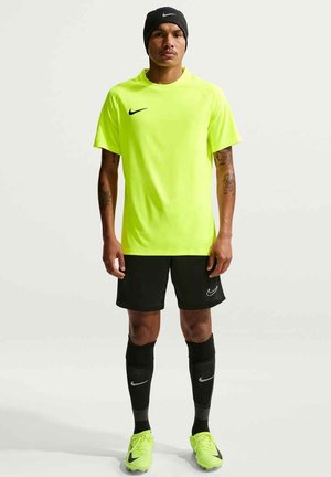 Sportowiec ubrany w neonowo-żółtą koszulkę Nike, czarne spodenki, czarne skarpetki, neonowo-żółte korki i czarną opaskę Nike na głowie, stojący na białym tle.