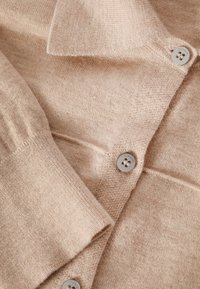 Gilet en tricot beige avec manches longues, col, et boutons visibles à quatre trous fixés le long de l'ouverture frontale.
