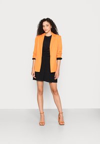 Blazer laranja sobre um vestido preto, de comprimento até o joelho, em tecido texturizado. Modelo usa sandálias de salto laranja, posando contra um fundo branco.