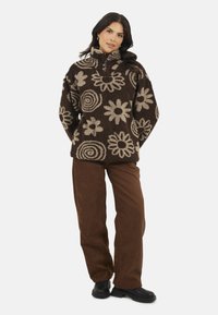 Donna che indossa un pullover di pile marrone con motivi floreali beige, pantaloni marroni e stivali neri, in piedi con le mani dietro la schiena.