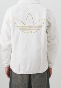 Bílá bunda se zlatě vyšitým logem s třemi listy a vodorovnými pruhy na zadní straně. Elastické manžety a lem, plátěná textura.