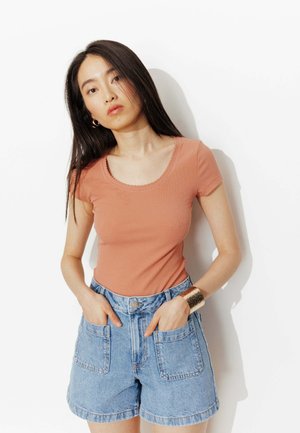 Jeune femme aux longs cheveux foncés portant un haut pêche à manches courtes et un short en jean taille haute, les mains dans les poches.