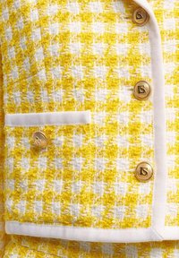 Žlutý a bílý tvídový kabát s kostkovaným vzorem, zlatými knoflíky s logy a bílou stužkou na kapse.