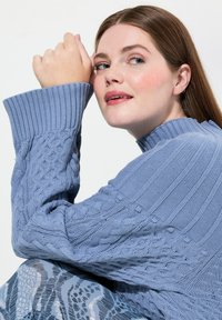 Ljusblå stickad tröja med ribbad krage och kabelstickat mönster, med långa ärmar. En par texturerade blå mönstrade byxor synliga.
