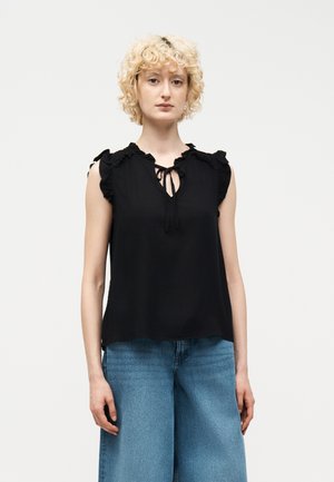 Γυναίκα με κοντά σγουρά ξανθά μαλλιά, φορώντας αμάνικη μαύρη μπλούζα με βολάν στους ώμους και μπλε τζιν παντελόνι με φαρδιά μπατζάκια, στέκεται μπροστά σε λευκό φόντο.