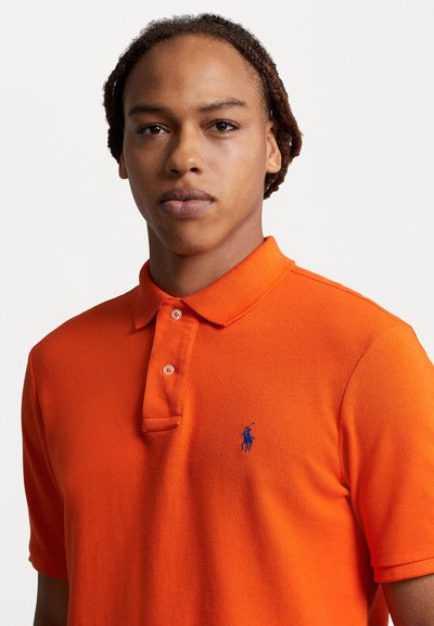 Oranž polo särk, mis on valmistatud tekstureeritud kangast; funktsioonid kraega kaelus, kahe nupuga nööpauk ja sinine tikitud logo rindkeres.