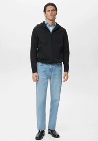 Sudadera negra con cremallera y capucha, combinada con jeans rectos de color azul claro. El modelo está de pie con las manos en los bolsillos, llevando zapatos negros.