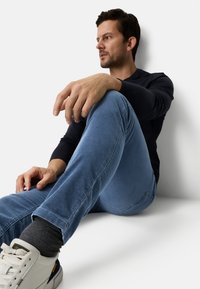 Blaue Cordhose, schwarzes langärmeliges Hemd und weiße Turnschuhe mit grauen Socken. Sitzende Pose, lässiges Outfit, glatte Texturen und schlichtes Design.