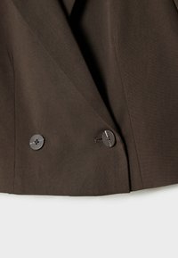 Gros plan sur un blazer marron foncé montrant le revers et deux boutons noirs sur la partie inférieure avant, sur fond blanc.