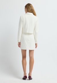 Blazer recortado texturizado blanco combinado con una falda mini blanca ajustada. El modelo lleva tacones granates. El conjunto cuenta con un collar y detalles de botones.