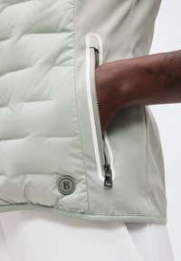 Χέρι με σκουρόχρωμο δέρμα που έχει τοποθετηθεί στην τσέπη ενός ανοιχτού ανοιχτό γκρι μπουφάν με επένδυση, με κυκλικό λογότυπο "B" κοντά στο τελείωμα.