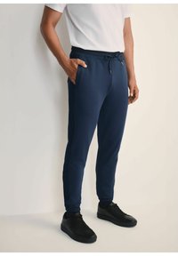 Granatowe spodnie dresowe wykonane z miękkiego materiału, z elastycznym pasem ze sznurkiem, bocznymi kieszeniami i zwężanymi mankietami. Widoczne czarne sneakersy.