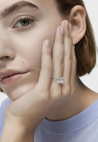 Anillo de plata con estrella y dos dijes de perlas, llevado en una mano apoyada contra el rostro. La modelo tiene el cabello corto y lleva un suéter azul claro.