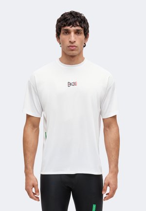Homme portant un t-shirt blanc uni à manches courtes avec un petit logo abstrait noir et rouge centré sur la poitrine.