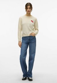 Maglione beige con una morbida texture e ricamo a forma di cuore rosso, abbinato a jeans blu a vita alta e scarpe nere con dettagli bianchi.