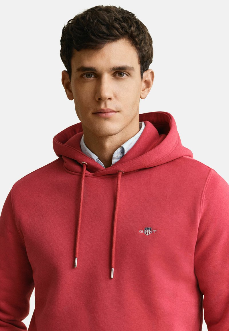 Junger Mann mit kurzen dunklen Haaren, der einen roten Kapuzenpullover über einem hellblauen Hemd mit Kragen trägt, schaut nach vorne mit neutralem Gesichtsausdruck.