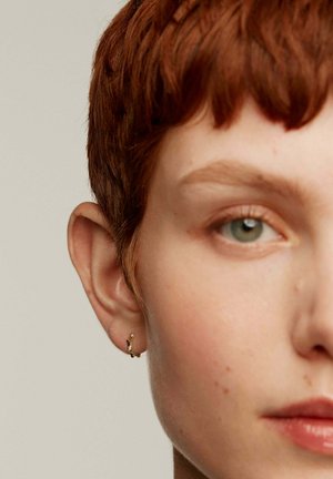 Primer plano de la cara de una persona con cabello corto y rojo. Las características notables incluyen un pendiente en forma de aro dorado y un ojo verde claro. Textura de piel suave.