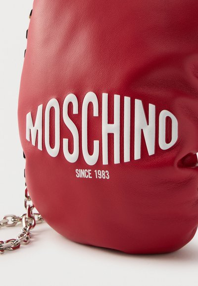 Κόκκινη δερμάτινη τσάντα με μεγάλο λευκό λογότυπο MOSCHINO και το κείμενο "Since 1983", με ασημένια αλυσίδα ως λουρί σε λευκό φόντο.