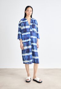 Rochie oversized bleu și alb cu tie-dye, cu decolteu adânc în V, mâneci trei sferturi și o buzunare frontal. Asortată cu sandale albe.