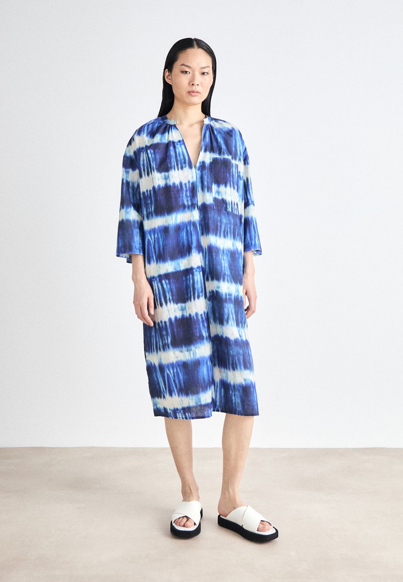 Rochie oversized bleu și alb cu tie-dye, cu decolteu adânc în V, mâneci trei sferturi și o buzunare frontal. Asortată cu sandale albe.