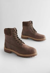 Botas de cuero marrón hasta el tobillo con un collar de ante, que presentan ojales de metal y una suela de goma con tacos para mayor tracción. Diseño robusto con detalles de costura.