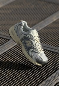 Sapatilha desportiva em cinza claro e branco com sobreposições em malha e sintético, repousando sobre uma superfície metálica texturizada.