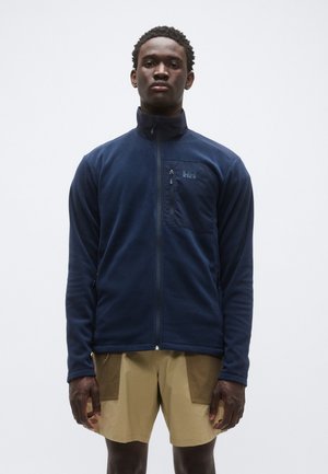 Veste polaire marine avec col montant, fermeture éclair frontale et poche poitrine. Elle présente une texture lisse et un logo ton sur ton sur la poche.