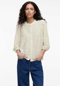 Light cream button-up blouse with a textured wavy pattern, feature collarless design and loose sleeves, paired with dark blue jeans.