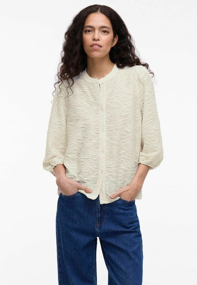Light cream button-up blouse with a textured wavy pattern, feature collarless design and loose sleeves, paired with dark blue jeans.