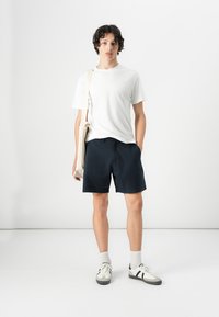 Un tricou alb cu mâneci scurte, pantaloni şort bleumarin şi adidaşi deschişi la culoare cu accente întunecate; modelul poartă o geantă de umăr bej.