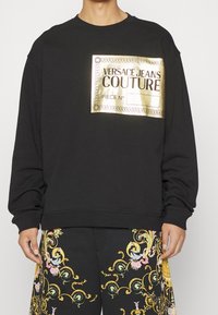 Svart sweatshirt med en guldbladsgrafiketikett med texten "VERSACE JEANS COUTURE". Långa ärmar och ribbade ärmslut. Mjuk tygstruktur.