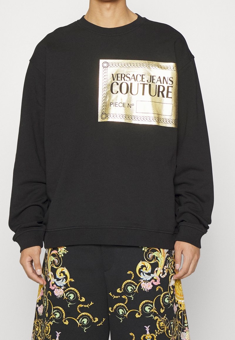 Svart sweatshirt med en guldbladsgrafiketikett med texten "VERSACE JEANS COUTURE". Långa ärmar och ribbade ärmslut. Mjuk tygstruktur.
