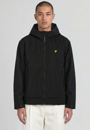 Uomo che indossa una giacca nera con cappuccio e zip con un piccolo logo giallo di un uccello sul petto e pantaloni bianchi, in piedi davanti a uno sfondo neutro.